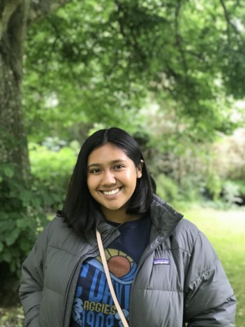 A young woman with shoulder-length dark brown hair wearing a blue UC Davis t-shirt and a grey puffer jacket smiles at the camera in front of a lush green background. 